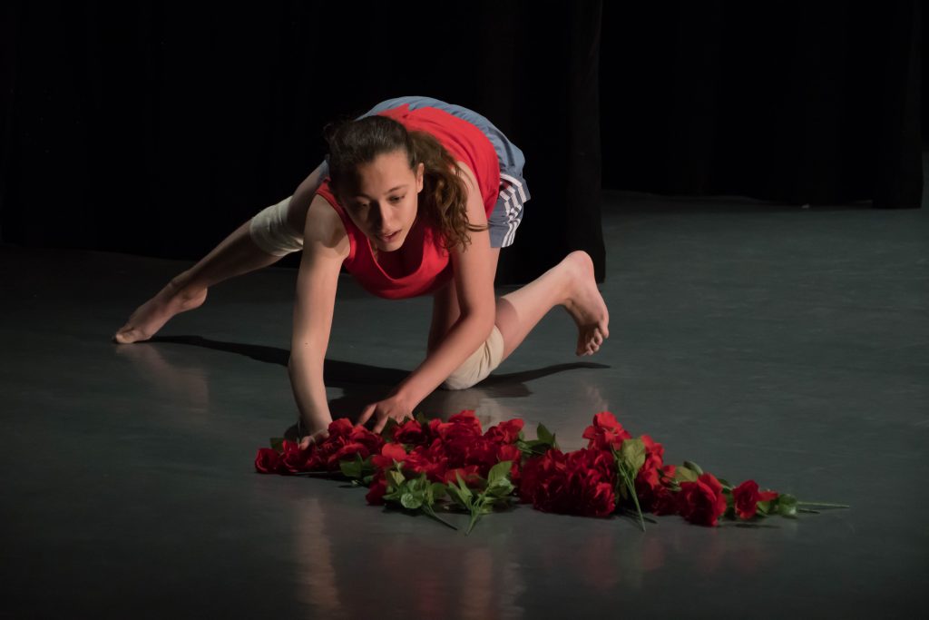Izzy is crouching with her left knee on the ground and her right leg outstretched. She is wearing beige kneepads, blue shorts, and a red top. She is grabbing at a pile of fake roses in front of her. The background of the image is a darkened stage. 
