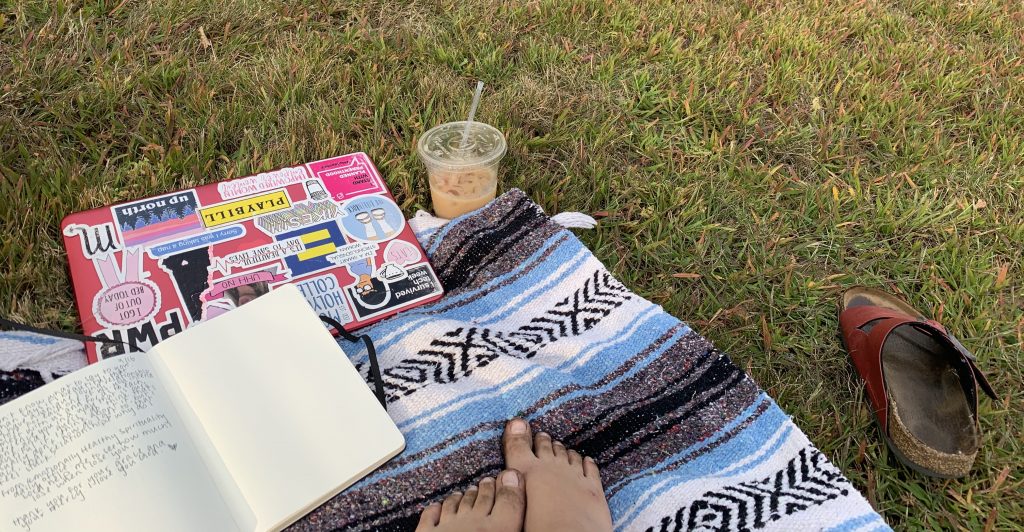 A patterned blanket is spread on green grass, a laptop covered in stickers, an open notebook with writing, a partially empty iced coffee, and two dirty bare feet resting on the
blanket. One red sandal is left alone next to the blanket.
