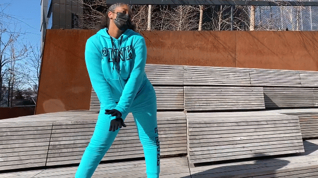 Gif shows Kayla, a Black woman with curly black hair, dancing outside on wooden slat steps. Kayla wears a light blue tracksuit with "PINK" written down the leg and across the chest, a black mask, black sneakers, and black gloves. Behind her is a rust-colored wall, barren trees, blue sky, and two NYC skyscrapers.