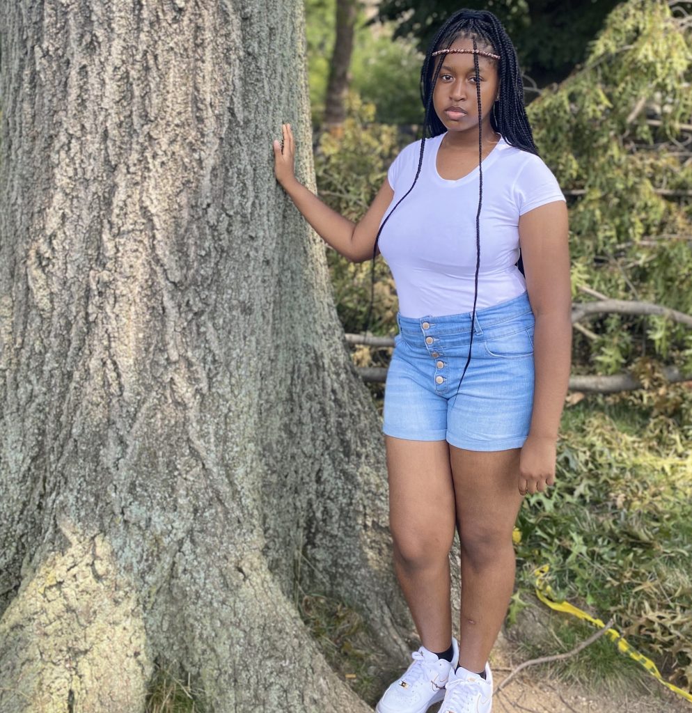 Picture shows Kayla, a Black woman with black box braids flooding down to her waist, facing the camera next to a tree with her right hand resting on its trunk. She wears a white t-shirt, blue jean shorts with gold buttons up the front, white Nike air forces, black ankle socks, and a skinny beaded headband across her forehead. Behind her is greenery in the form of standing and fallen trees, as well as a glimpse of caution tape strewn across the ground.