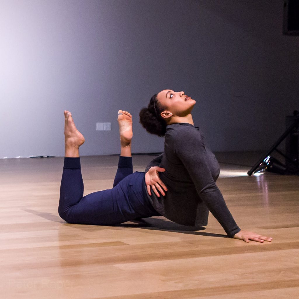 Photo shows Olivia, a Black woman with curly brown hair, posing mid slide in a dance studio space with a wooden floor and white wall. She thoughtfully gazes towards the ceiling with her back arched. Her lower legs are bent at near ninety degree angles with her feet pointing upward as her right palm presses into the floor for support and her left arm reaches across her lower back, palm facing open. Olivia wears a gray long sleeve shirt and navy blue athletic pants.
