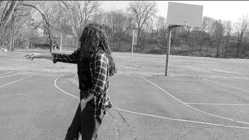 a black and white still from Aggie's capstone. They are on an empty basketball court , facing to the left with one arm extended in from of them. Their long hair obscures their face completely.