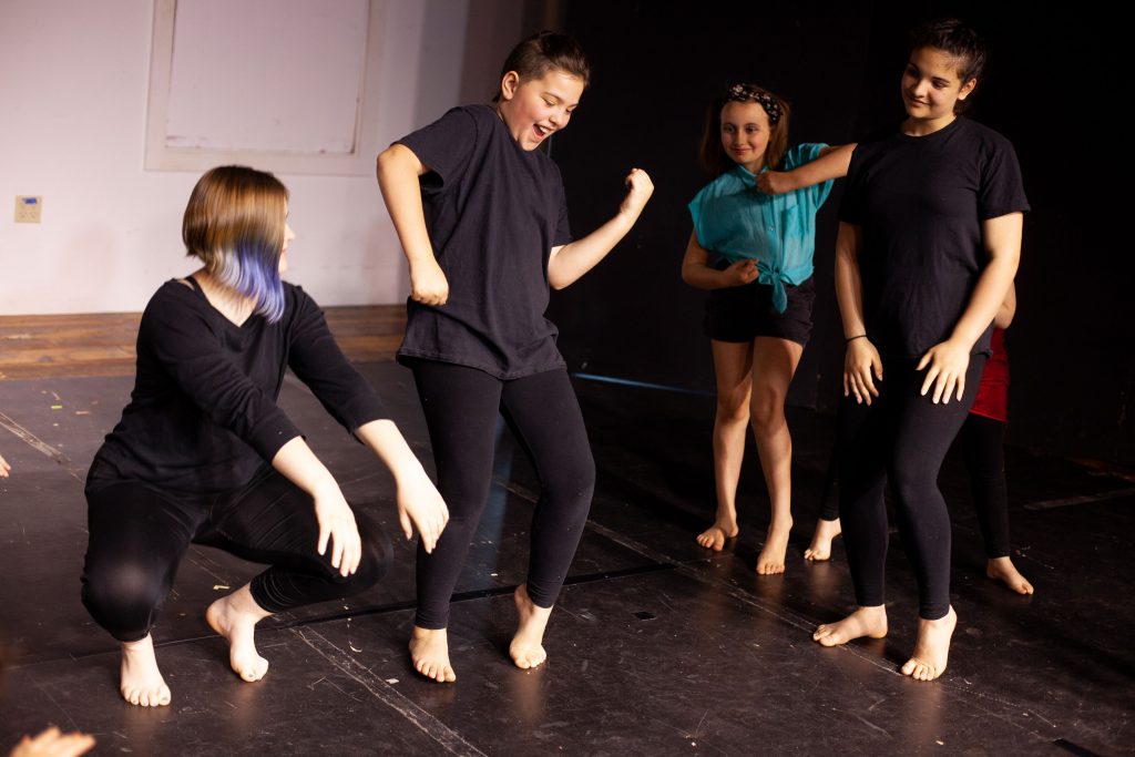 Photo shows four adolescents (aged 10 - 12) dancing casually on a black marley floor. Youth are looking at each other, smiling, and appear in various different positions.