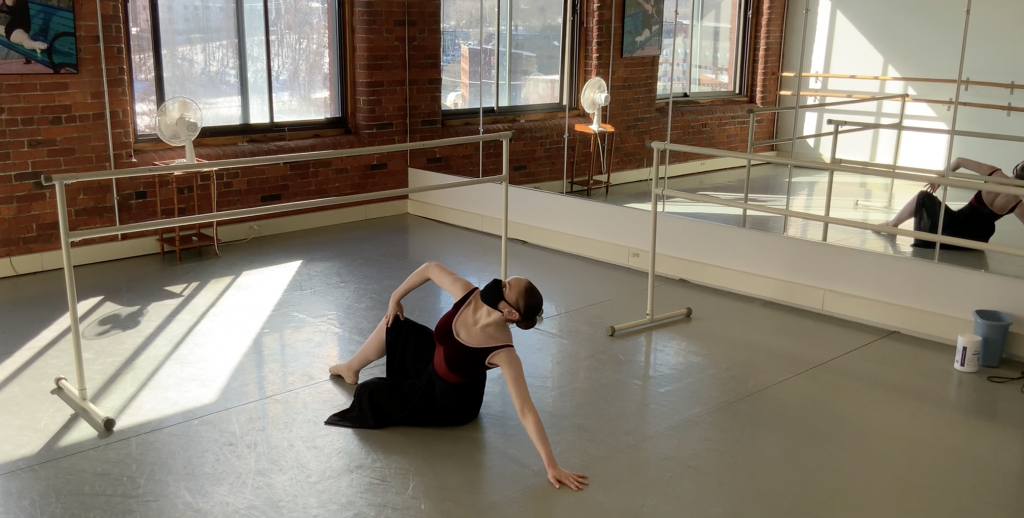 Snapshot of Hannah during rehearsal for Rose Flachs’ choreography. Hannah is sitting on the ground in a ballet studio in a strong position with one hand on her knee and the other reaching on the ground. She wears a dark red leotard, pink tights, ballet slippers, a long black skirt and a black face mask. Next to and behind her are ballet barres. 
