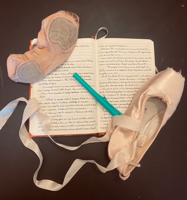 A ballet shoe, a pointe shoe, and a blue pen rest on an open notebook covered in handwriting. The background of the photo is black.