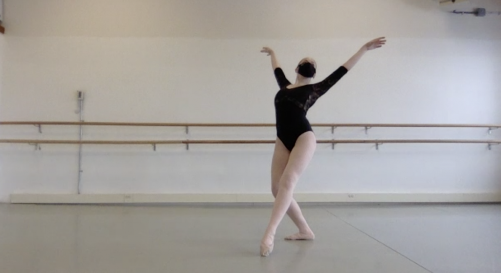 Snapshot of Hannah during Joseph Jefferies’ rehearsal. She is in a ballet studio with a ballet barre behind her that is attached to a white wall. Hannah is in croise devant in plie with her arms in fifth allongé. She wears a long sleeved black leotard, pink tights, ballet slippers, and a black face mask.  
