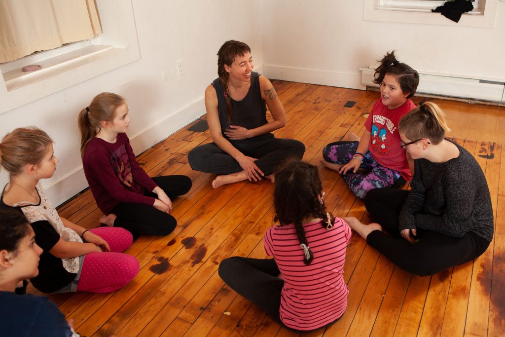 
Photo shows Anna Hendricks sitting cross legged in a circle alongside 7 adolescent youth (ages 10 - 12). They have bare feet, are dressed in casual clothing and are on a wooden dance floor. Anna is smiling and looking at a student who is speaking. She has her hand placed on her stomach.
