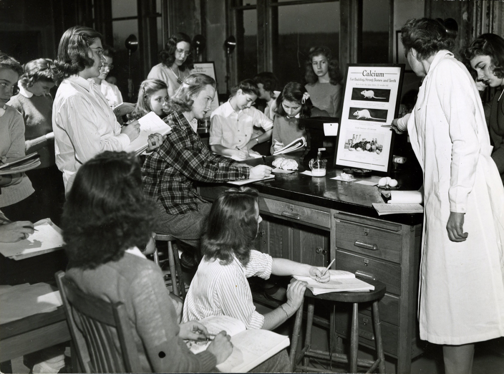 Female students sit around a desk, taking notes on a presentation. A female teacher in a white lab coat point to a presentation board with mice on it. The classroom has large windows and wooden stools and desks.