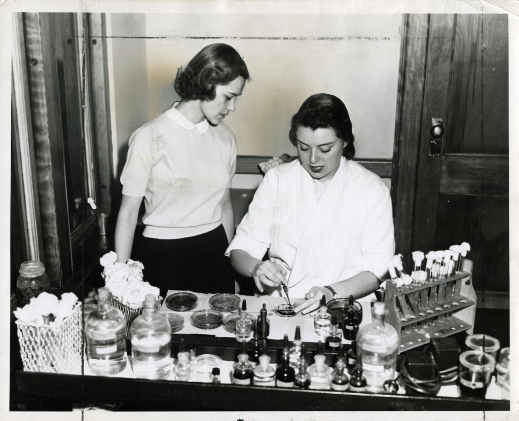 Two young female students using chemistry equipment in a lab. Glass jars of chemicals and a rack of test tubes are across the desk. A sitting student is applying chemicals in a petri dish while wearing a lab coat. Another student is standing to her left, looking at the dish, and is wearing a white collared shirt.