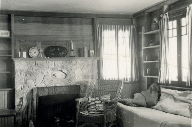 A living room with wooden walls, a stone fireplace, and curtained windows. A wicker chair with a pillow sits in front of the fireplace, and a couch sits to the right. The fireplace's mantle is decorated.