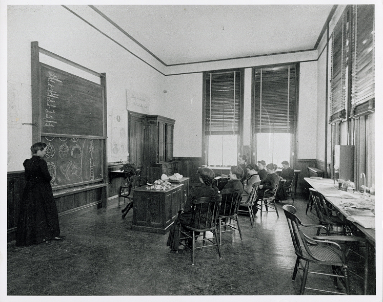 A classroom with students sitting in wooden chairs, facing a chalkboard. A teacher stands next to the chalkboard, which has scientific drawings on it. The classroom has wooden floors, tall, white walls, and sunlit windows.