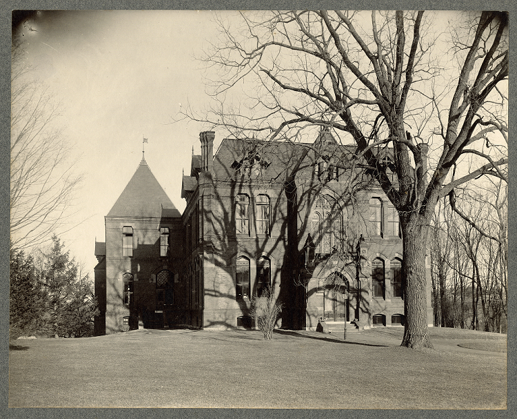 A two story brick building with arched windows. There is a similar brick annex attached to the left, with a peaked roof. A large, leafless tree sits in front of the building.