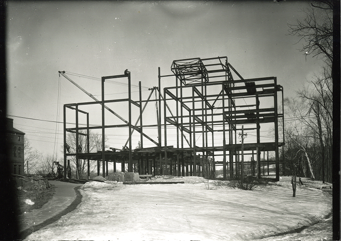 A metal beam structure during the construction of Clapp Building. There is snow on the ground. A crane is behind the structure.