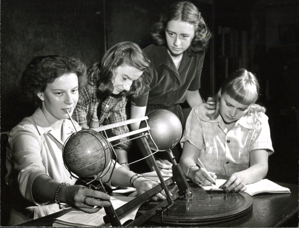 Four students gather around a model of the solar system. Two students look down at papers as they take notes, another student looks at the device, and the last student is both looking at and touching the device.