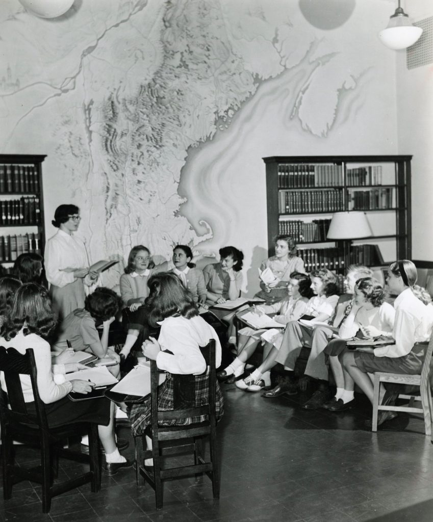 Students sit in chairs in a circle, while they listen to a professor lecture. The room has two large bookcases, sat against a wall with a marble pattern.