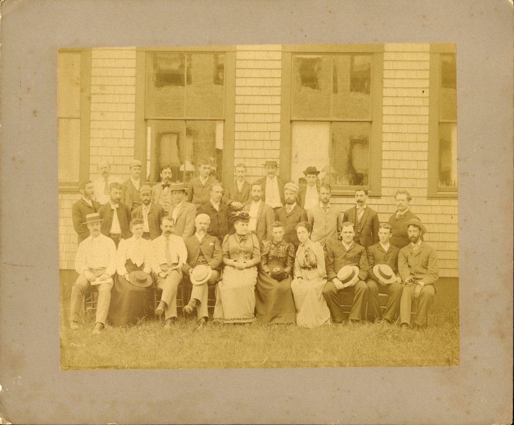 A group of men and women pose together in three rows, with the first row seated and the last two standing. They're outside on grass, posed in front of a white shutter building with large windows.