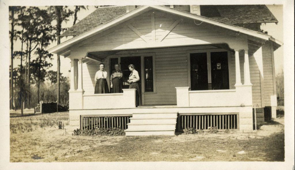 Three women stand together on the porch of a house. The house is white with a covered porch, and stairs leading up. Trees are in the background.