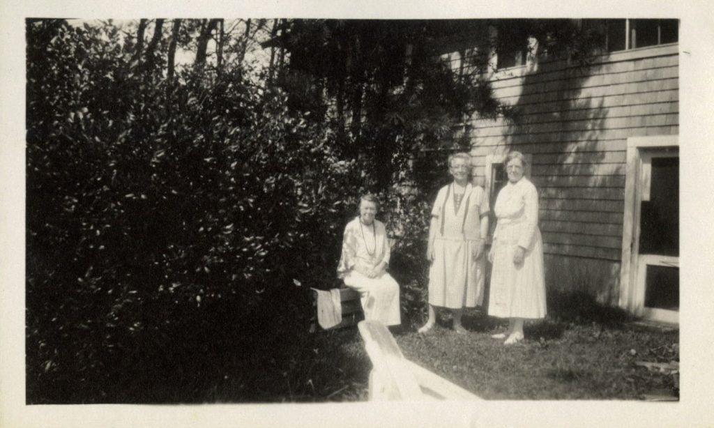 Three women pose in a backyard. They all wear white dresses, and two stand while one sits. There are trees and a shuttered house behind them.