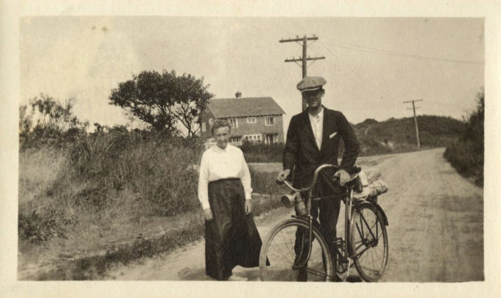 A woman and a man stand in a road. The man holds a bike. There are trees, a house, and power lines in the background.