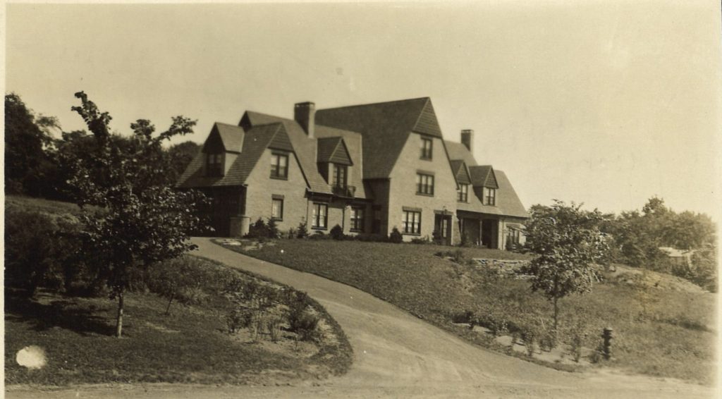 A large house sits on a hill. A driveway leads up to it, with multiple trees in the grass yard.