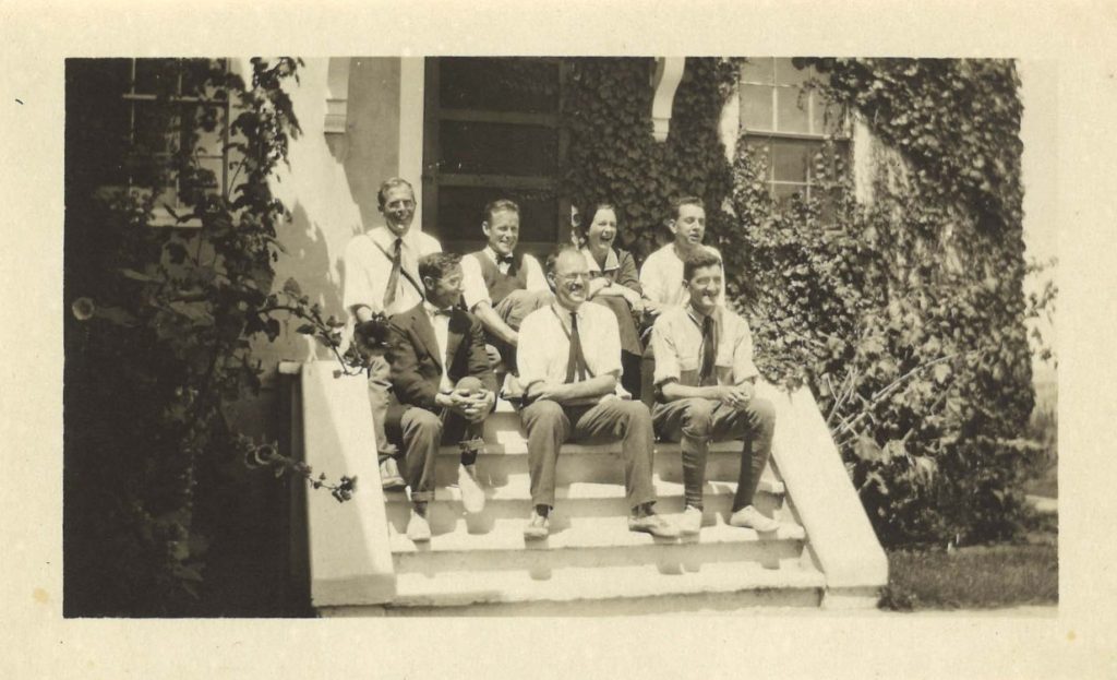 Six men and one woman sit on steps outside a home, smiling at the camera. Ivy covers the wall behind them, as well as shrubbery next to them.