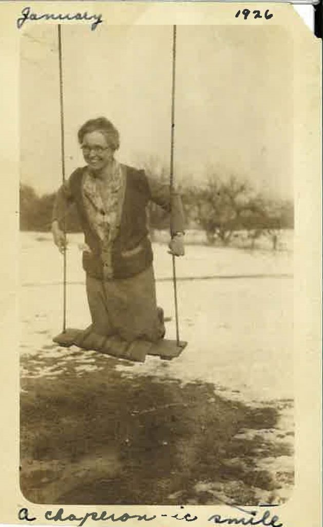 A woman smiles as she swings on her knees on an outdoor wooden swing. There is snow on the ground and trees in the distance.