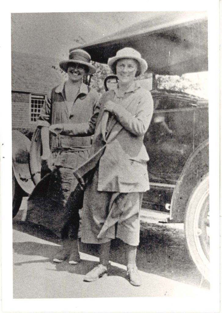 Two women stand in front of a car. They both wear hats and coats, and smile at the camera.