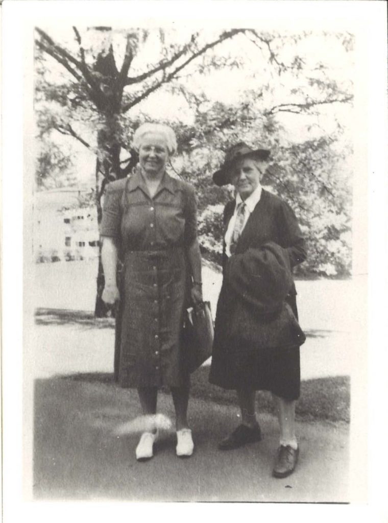 Two women pose for a photo standing in front of a tree. They both wear long dresses, while one wears a hat.