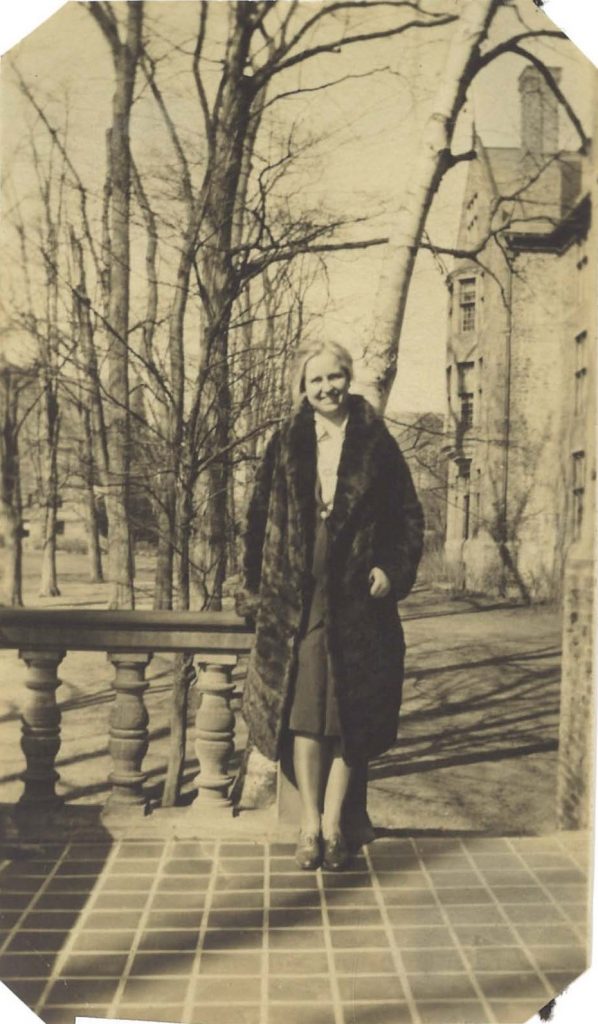 A woman in a fur coat poses outside a brick building, on a patio with a stone fence. The trees behind her are bare.