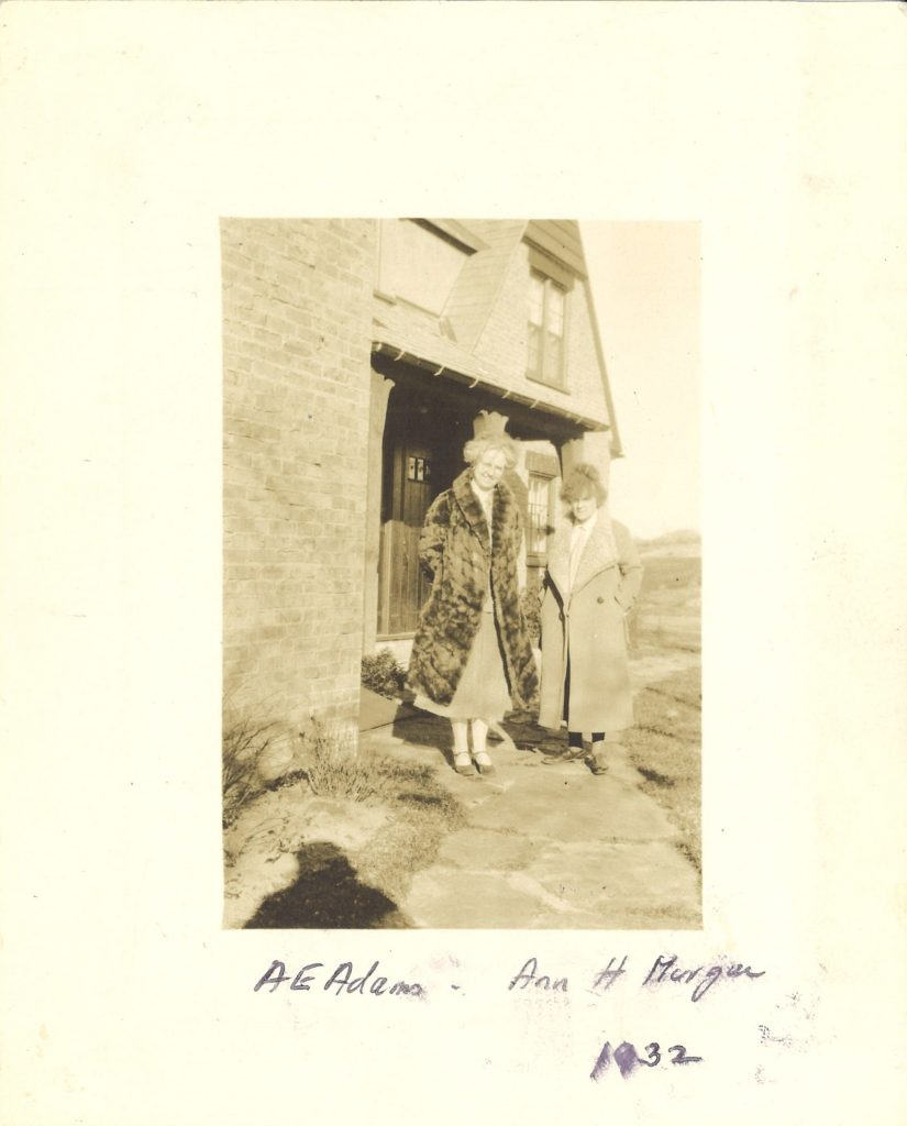 Two women pose for a photo outside a brick house. They both wear long coats.