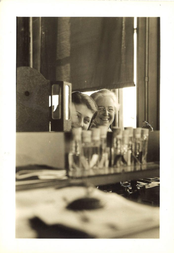 An older woman and younger student smile for a photo, partially obstructed by lab equipment, including glass bottles.