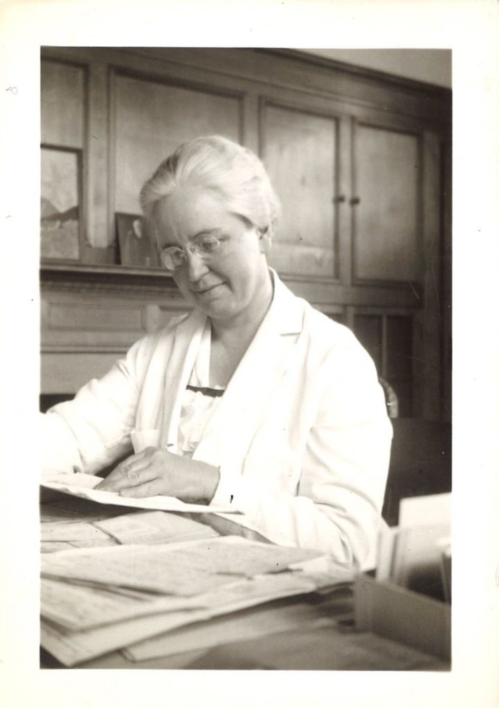 An older woman in a lab coat sits at a desk, reading notes.