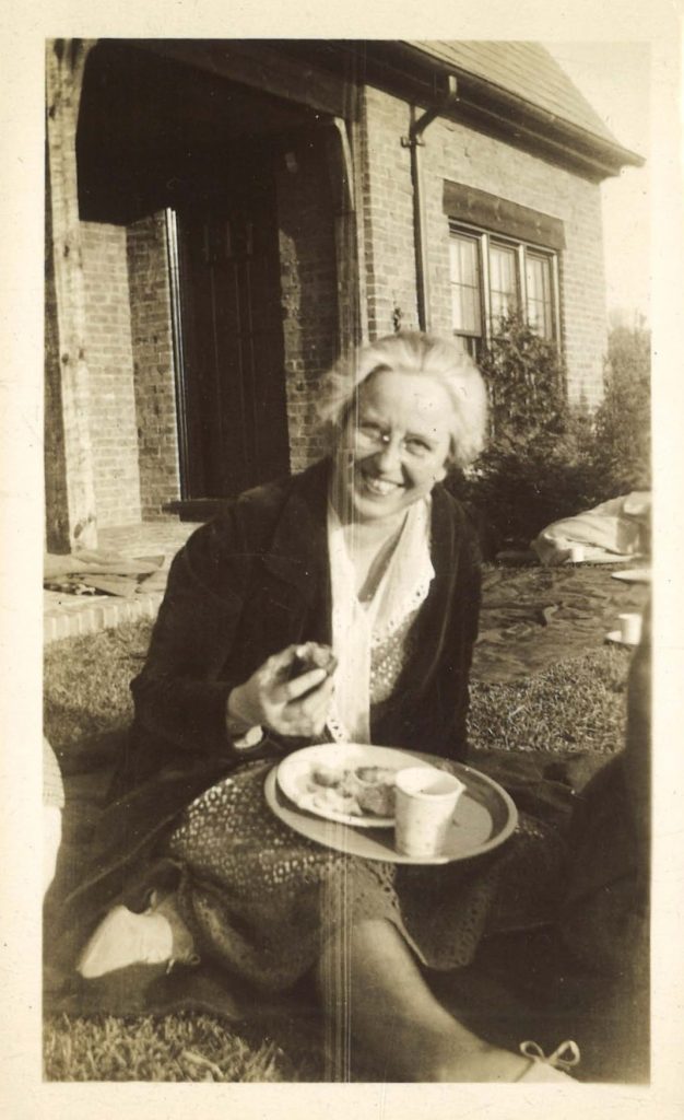 Woman sits in grass, eating food off of a plate. She smiles at the camera. There is a brick house behind her.