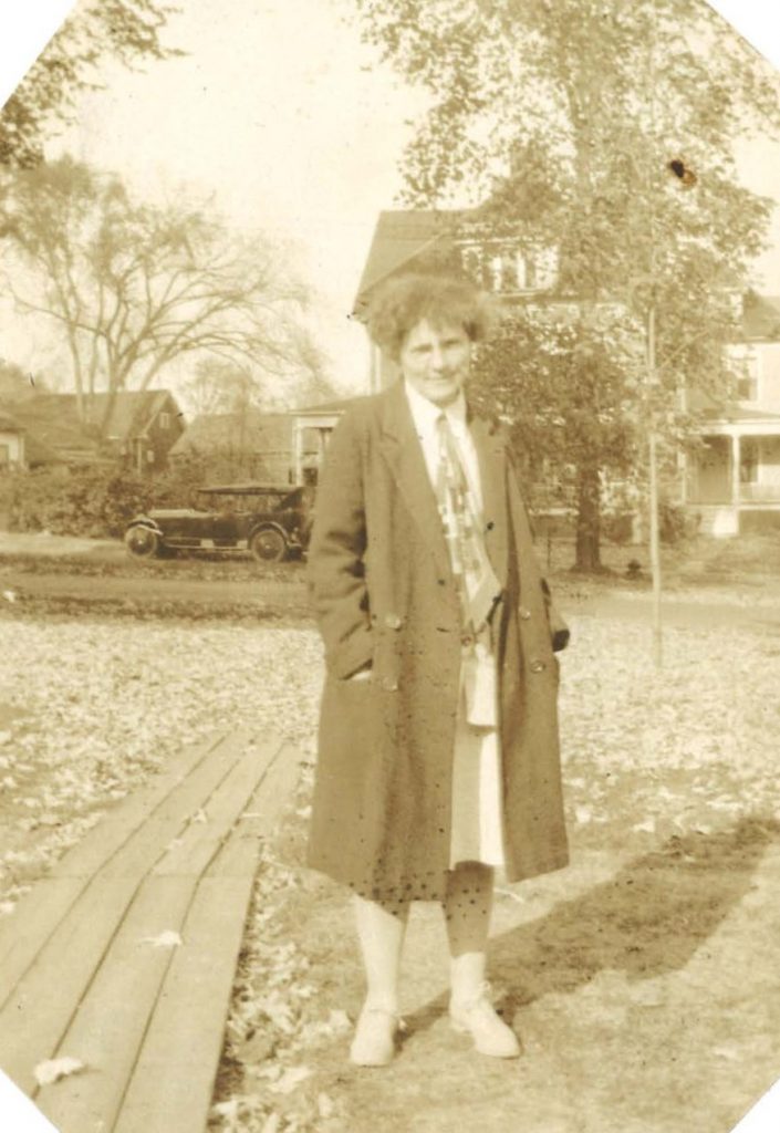 A woman poses outside in the grass, in front of a house, car, and tree. She wears a long coat.