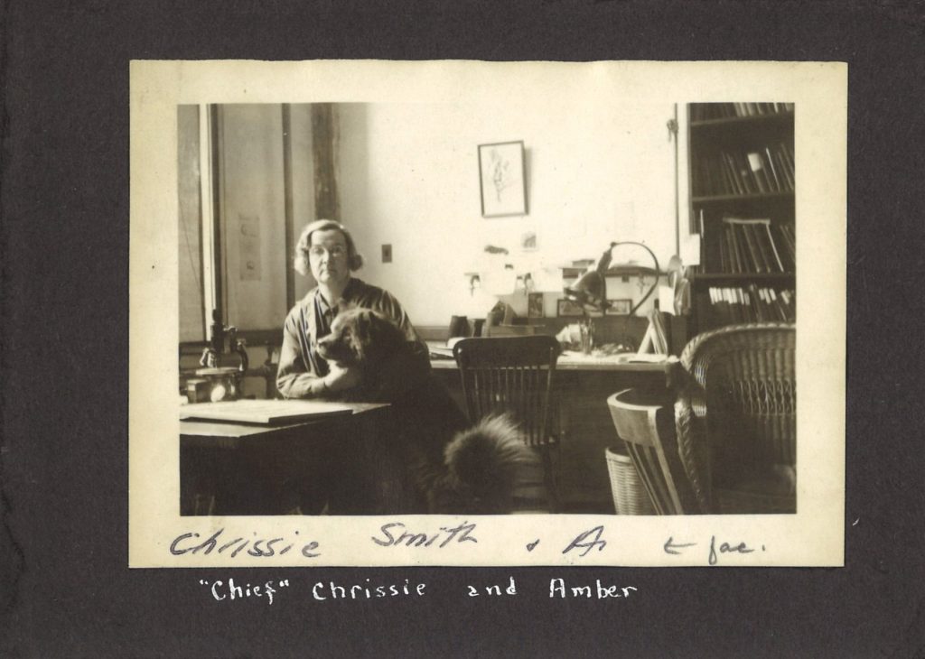 A woman sits at a table in an office, posing with a large, fluffy dog. Caption on photo reads "'Chief' Chrissie and Amber."