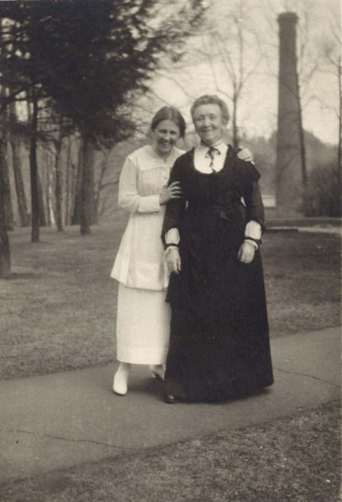 A smiling, young Ann Morgan in a white dress poses with her hands on the arm and shoulder of an older Cornelia M. Clapp. Clapp wears a dark, collared dress. They stand outside on pavement, with trees and a tall, narrow building behind them.