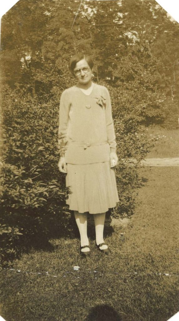 A woman poses outside in front of a bush. She swears a sweater and skirt.