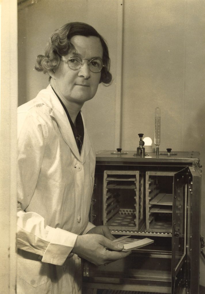 An elderly woman wears a white lab coat while she holds a microscope slide.