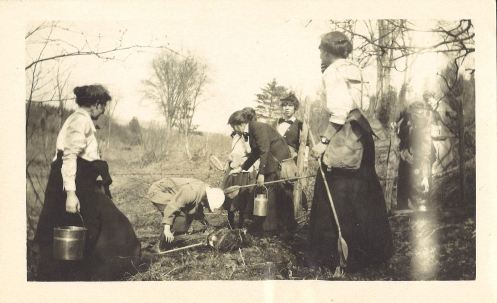 Seven female students in a field digging through the dirt in search of specimen. They are wearing white button ups and black skirts, and hold shovels and buckets.