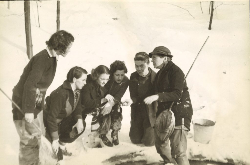 Five students and a professor huddle around an ice hold with nets and buckets. There is heavy snow around.