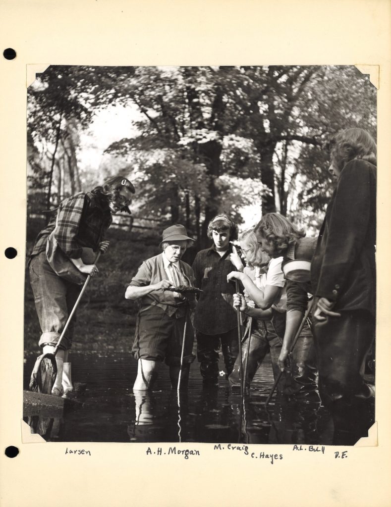 Five students and one professor stand in a body of water. They are holding nets and looking at the specimen the professor holds.