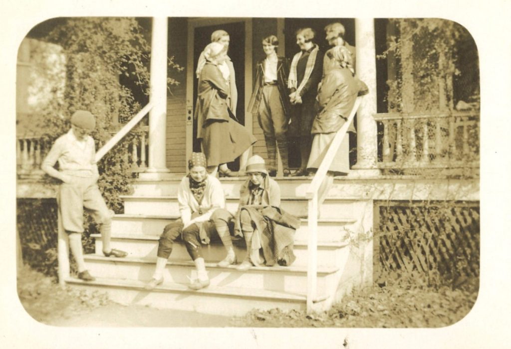 Eight students stand and talk on a house's outside staircase, while two students sit and talk on the staircase. There are plants around the staircase, and it leads to a porch.