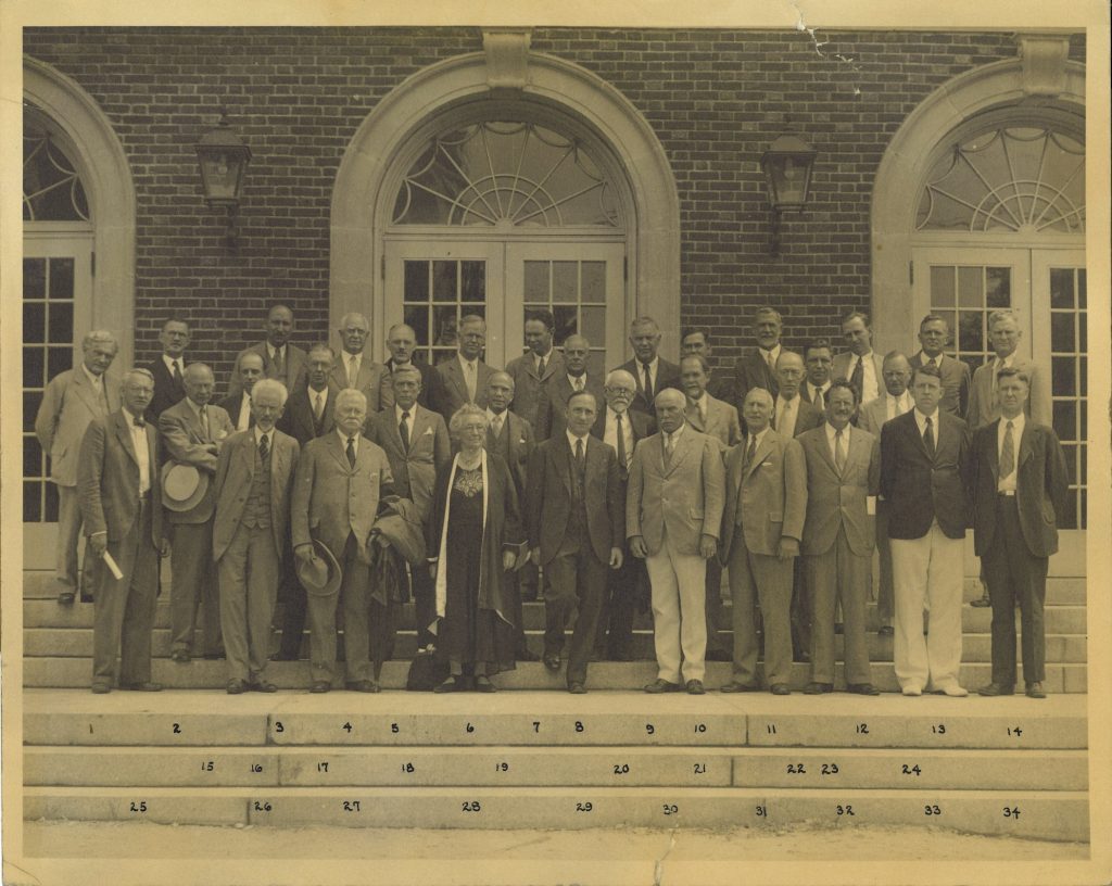 A group photo of men in suits, with one woman in the front. They are standing on stone steps, in front of a brick building with arched windows.