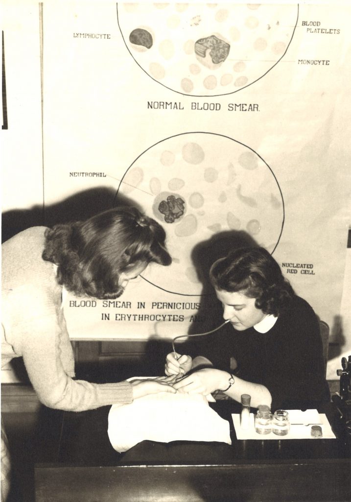 One student sitting pricks the finger of another student standing in a blood lab. There is a poster behind them showing different types of blood smears.