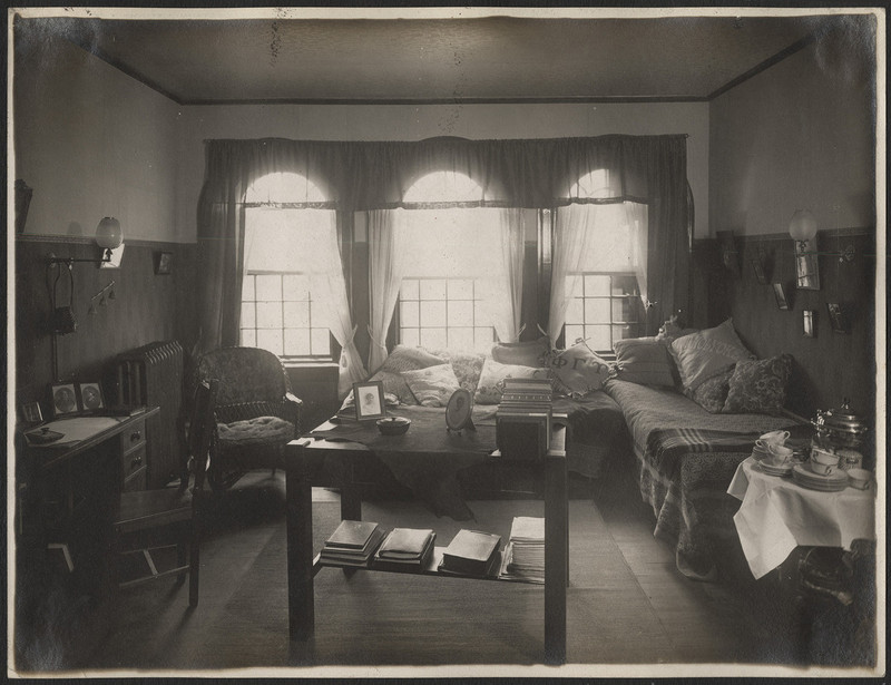 Photograph of interior of dorm room, includes table in the center with photographs, notebooks, and table with tea set. The back wall has three large windows with curtains and sunlight coming through.