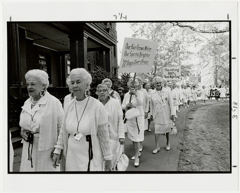 A line of women dressed in all white walk along a sidewalk outside a dormitory. Two are holding signs.