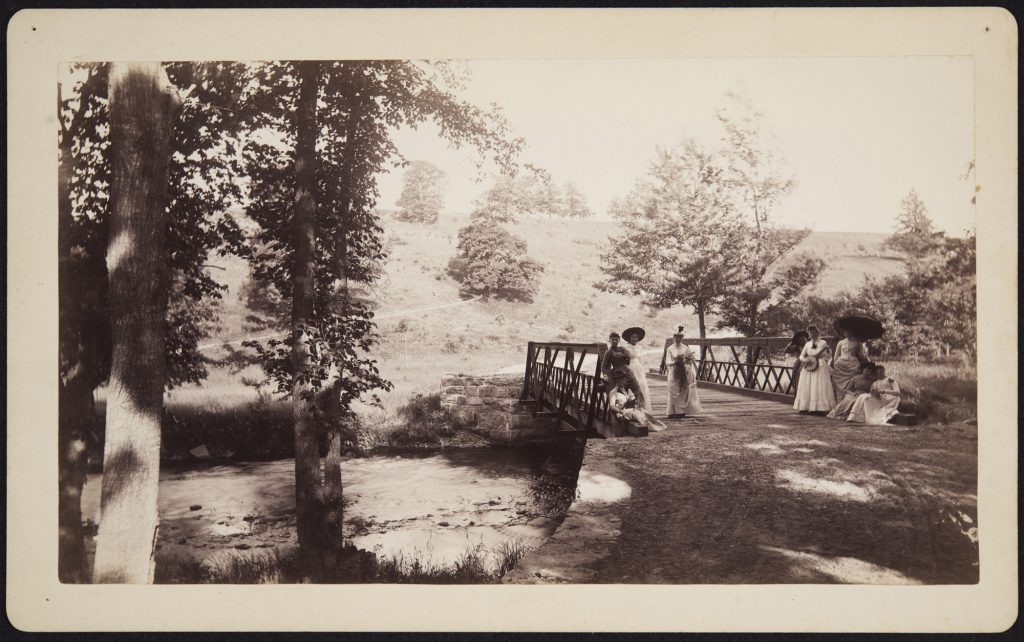 Women dressed in dresses and hats, one holding a parasol, stand on a bridge with a hill behind them.