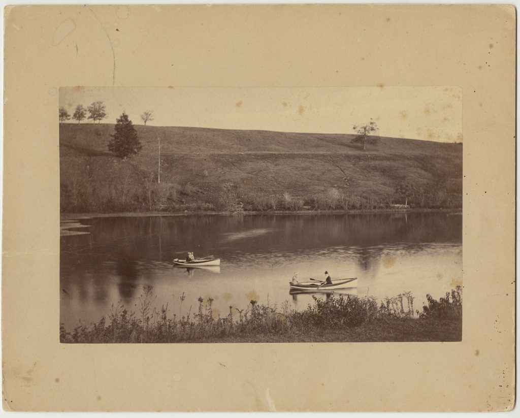 Two canoes, one person in each, sit on the water of a lake with a hill behind them.