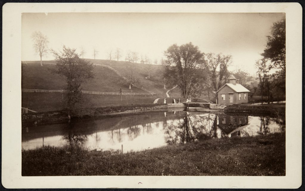 A small building sits beside a lake with a hill behind it. The hill has a few trees, and lines of fencing.