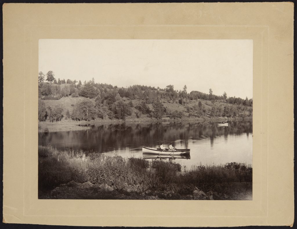 Two people row a canoe on a lake with a tree dotted hill behind them.